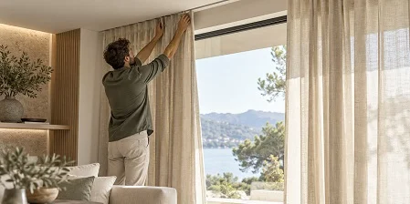 Man hanging beige linen curtains in a room overlooking a lake