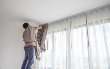 Man on a ladder hanging linen curtains over sheer white window
